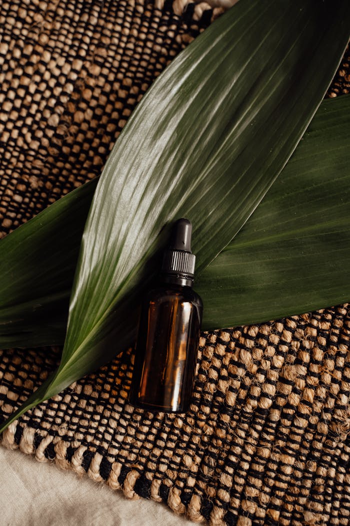 Brown glass dropper bottle on woven mat with green leaves, top view.